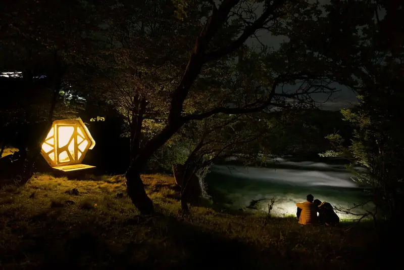 Un micro-refugio encendido de noche junto al río Cochrane, con dos personas sentadas en la orilla