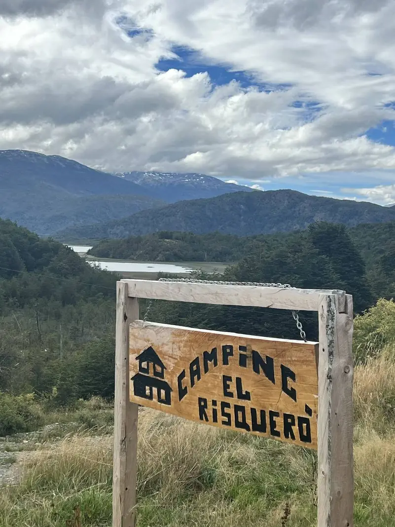 El letrero del Camping El Risquero, con el valle y el lago detrás
